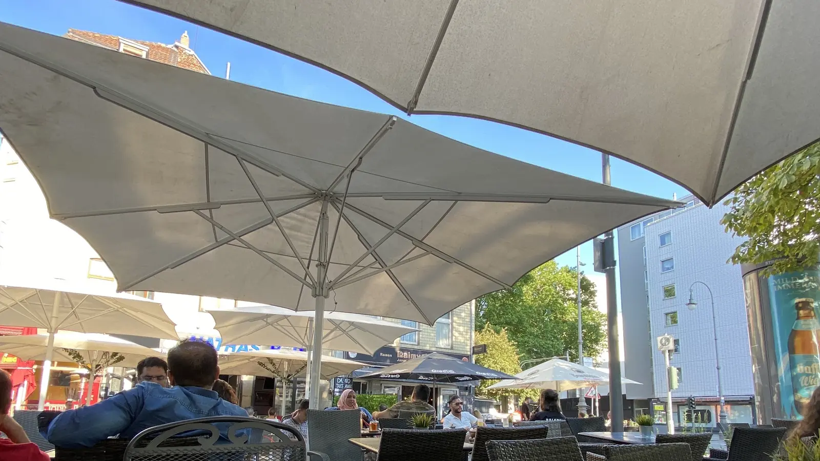 Die Terrasse bei Tageslicht: helle Sonnenschirme ueber Korbstuehlen, dahinter die Friesenstrasse mit Baeumen und Haeuserfront